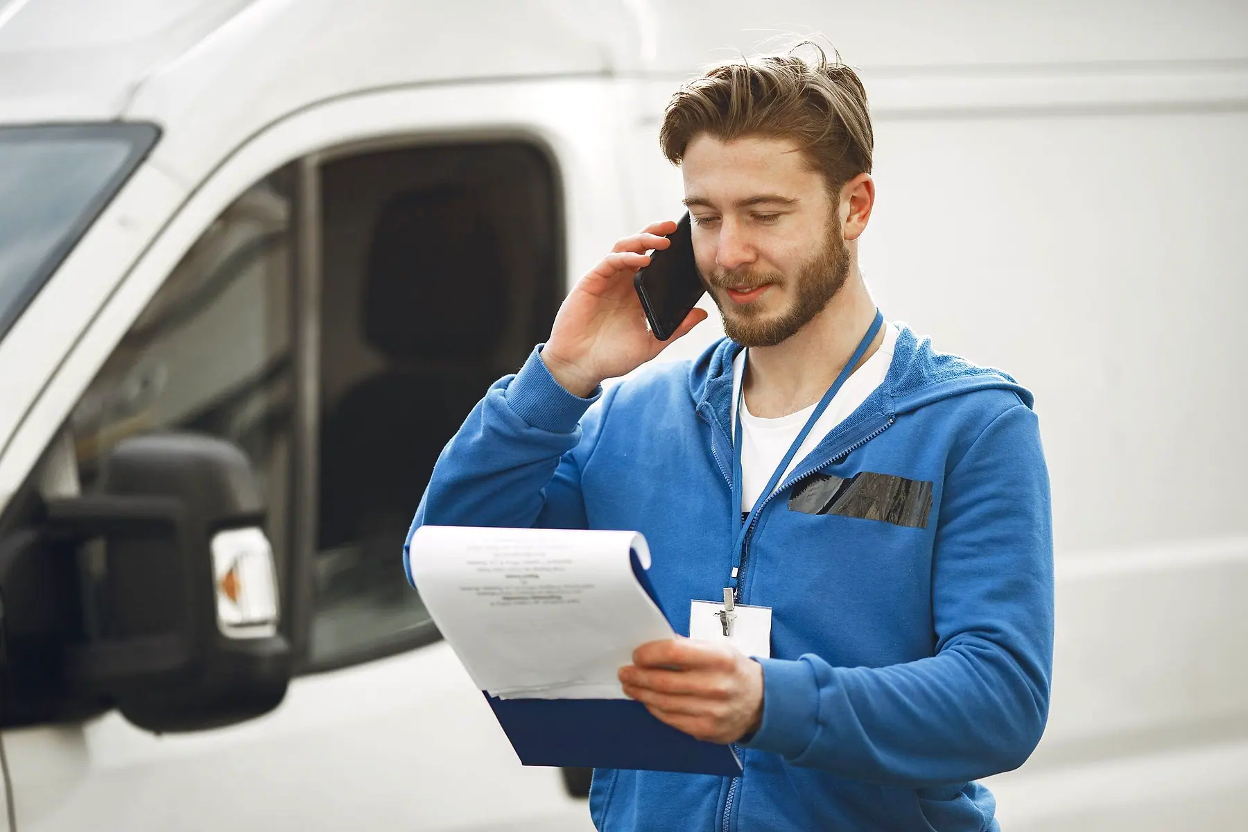 A tradesman beside his van taking a call while checking a job sheet on a clipboard