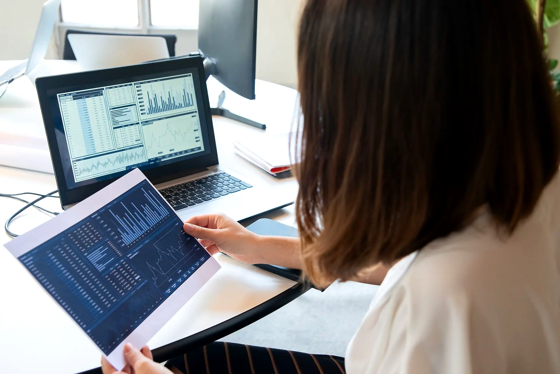 A woman comparing charts on a tablet and a laptop at her desk