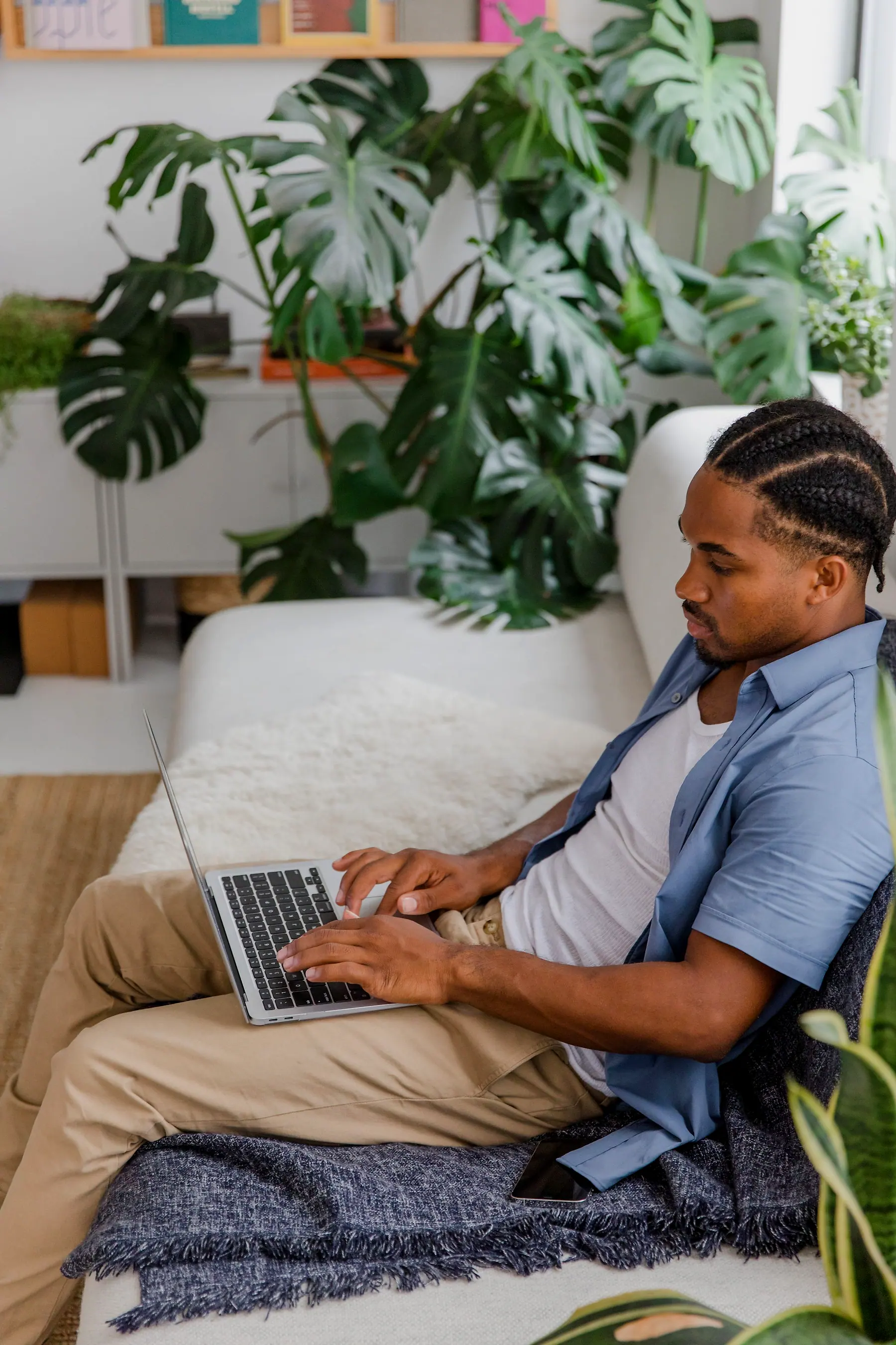 A man with braided hair typing on a laptop on a sofa in a plant-filled living room
